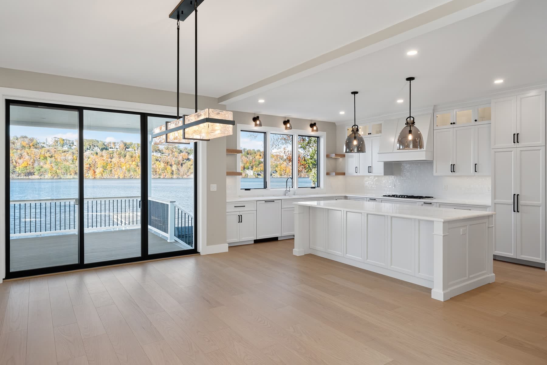 Open white kitchen at 148 North Lake Shore with a large island, bronze pendant lighting and sliding doors to the lake-view deck — Bulldog Enterprises.