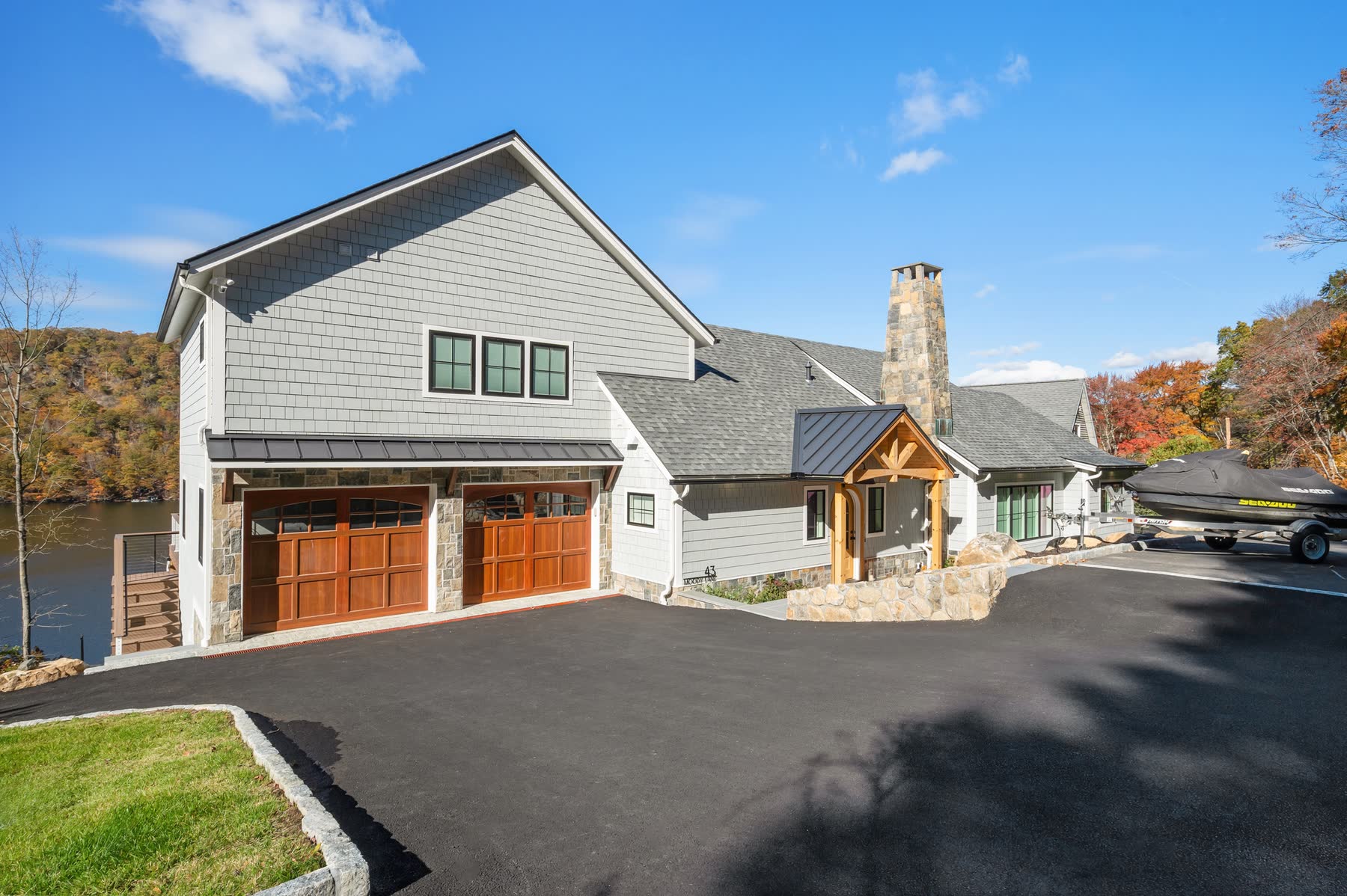 Front of the gray cedar-shingle home at 43 Moody Lane with carriage-style wood garage doors, a stone chimney and a lakeside drive on Candlewood Lake — Bulldog Enterprises.