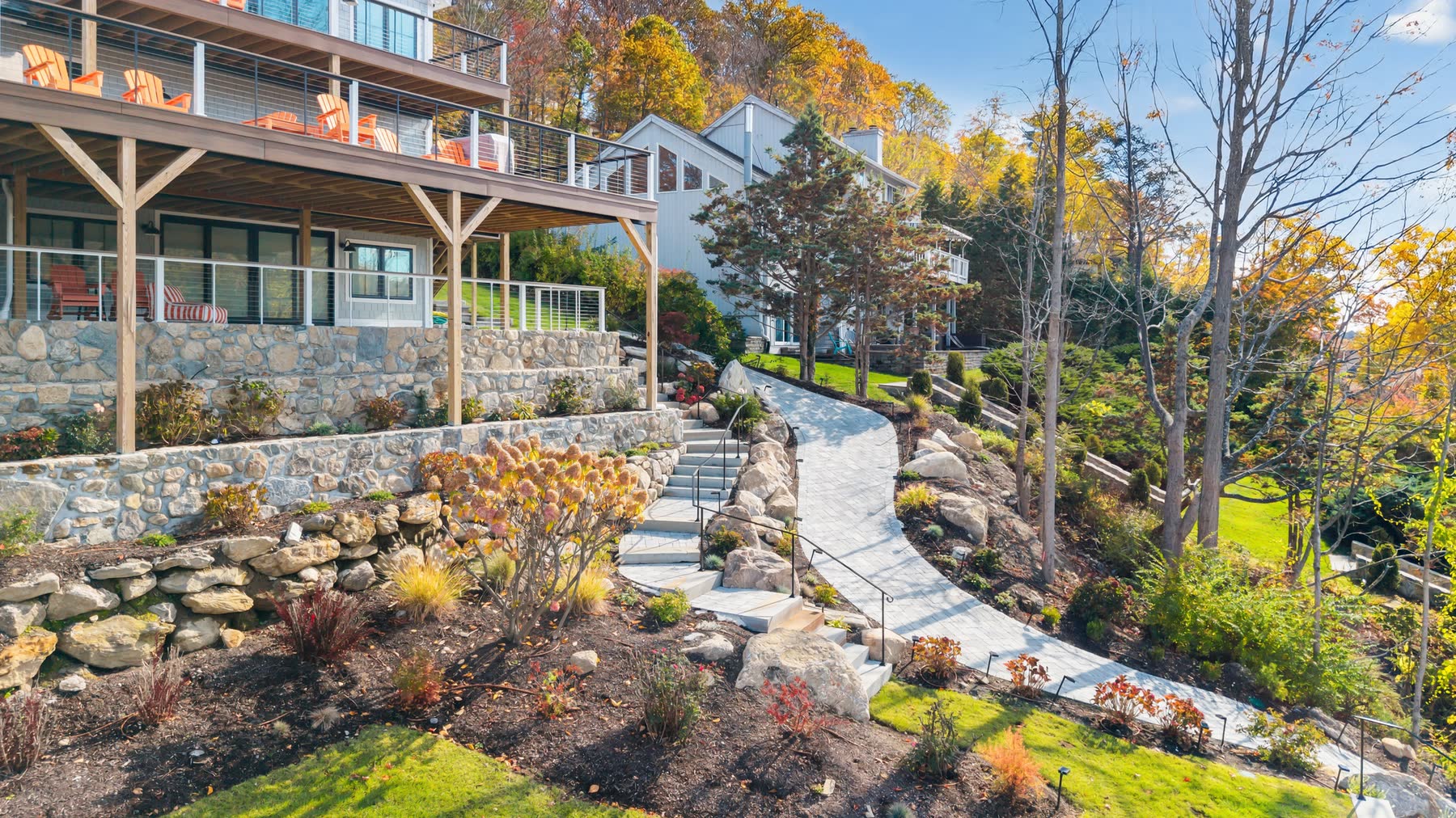 Elevated view of the rear of 43 Moody Lane — a covered porch with Adirondack chairs above stone retaining walls and a bluestone path stepping down to the lake — Bulldog Enterprises.