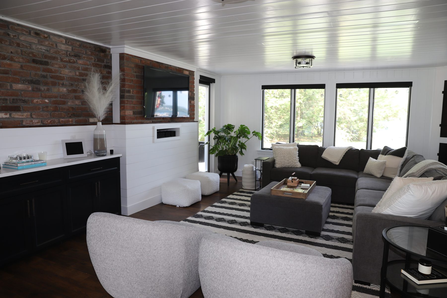 Living room with an exposed brick accent wall, mounted TV, shiplap plank ceiling and a sectional grouped on a striped rug - Sunset Drive, a renovated lake home on Candlewood Lake