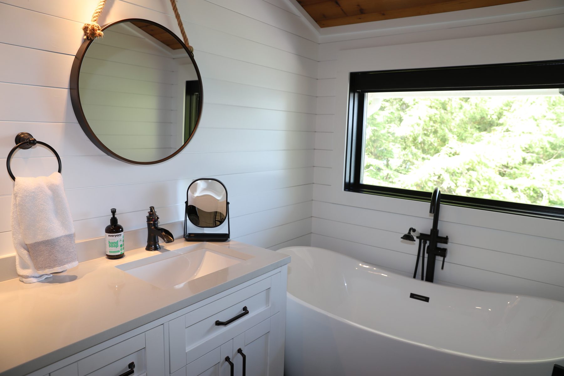 Spa-style bath with a freestanding soaking tub, shiplap walls, a rope-hung round mirror and a black-framed window - Sunset Drive, a renovated lake home on Candlewood Lake