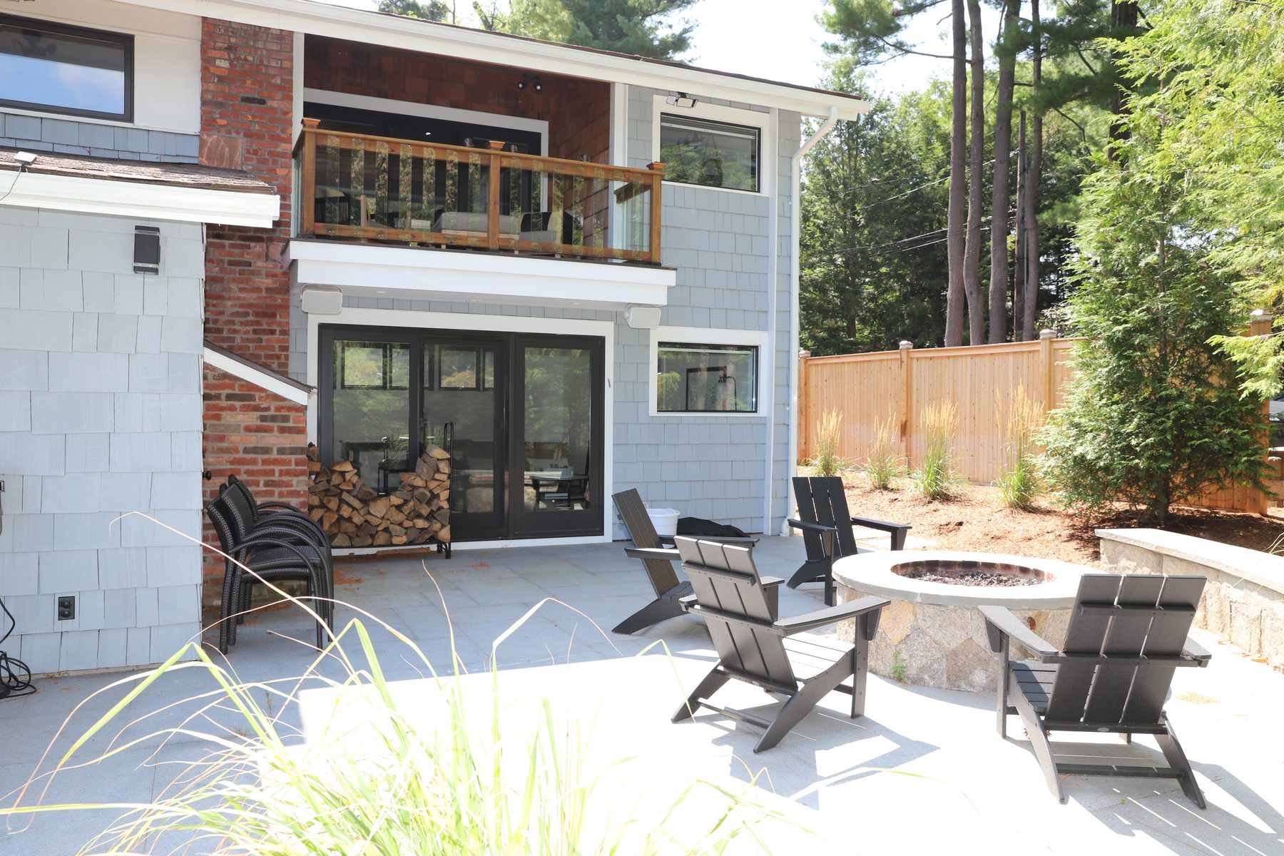Rear courtyard with a stone fire-pit and Adirondack chairs below a brick-and-shingle facade and second-floor balcony - Sunset Drive, a renovated lake home on Candlewood Lake