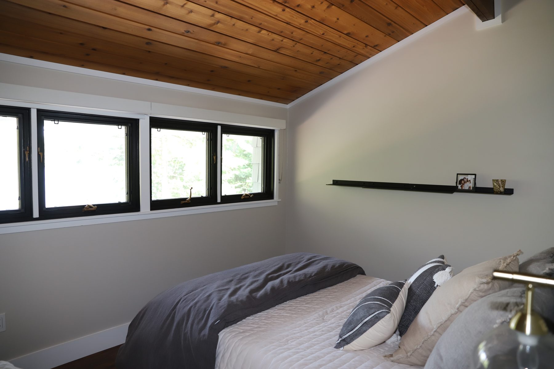 Bedroom under a warm wood-plank vaulted ceiling with a band of black-framed ribbon windows and a floating shelf - Sunset Drive, a renovated lake home on Candlewood Lake