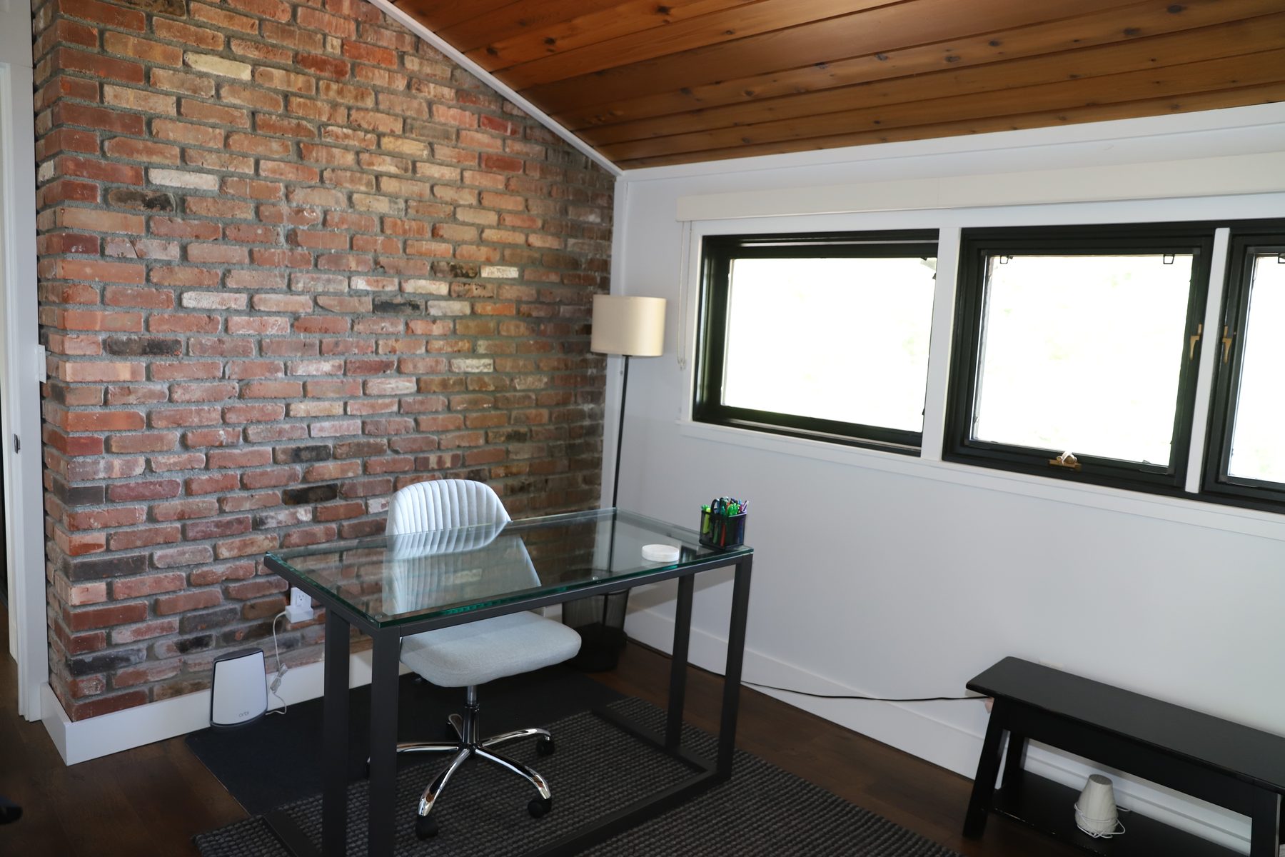 Home office with a full exposed-brick wall, glass-top desk and wood-plank ceiling beside black-trimmed windows - Sunset Drive, a renovated lake home on Candlewood Lake