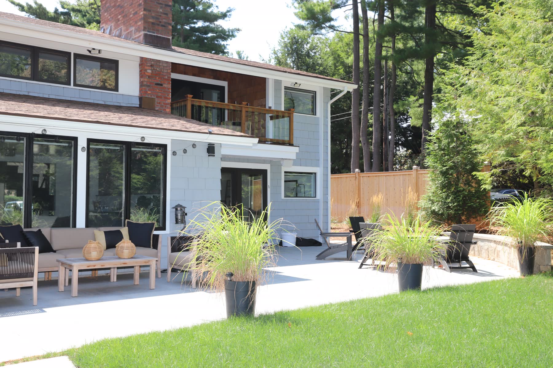 Rear of the mid-century-modern home at Sunset Drive with gray board siding, a brick chimney, an upper balcony and a bluestone patio set for outdoor living — renovation by Bulldog Enterprises.