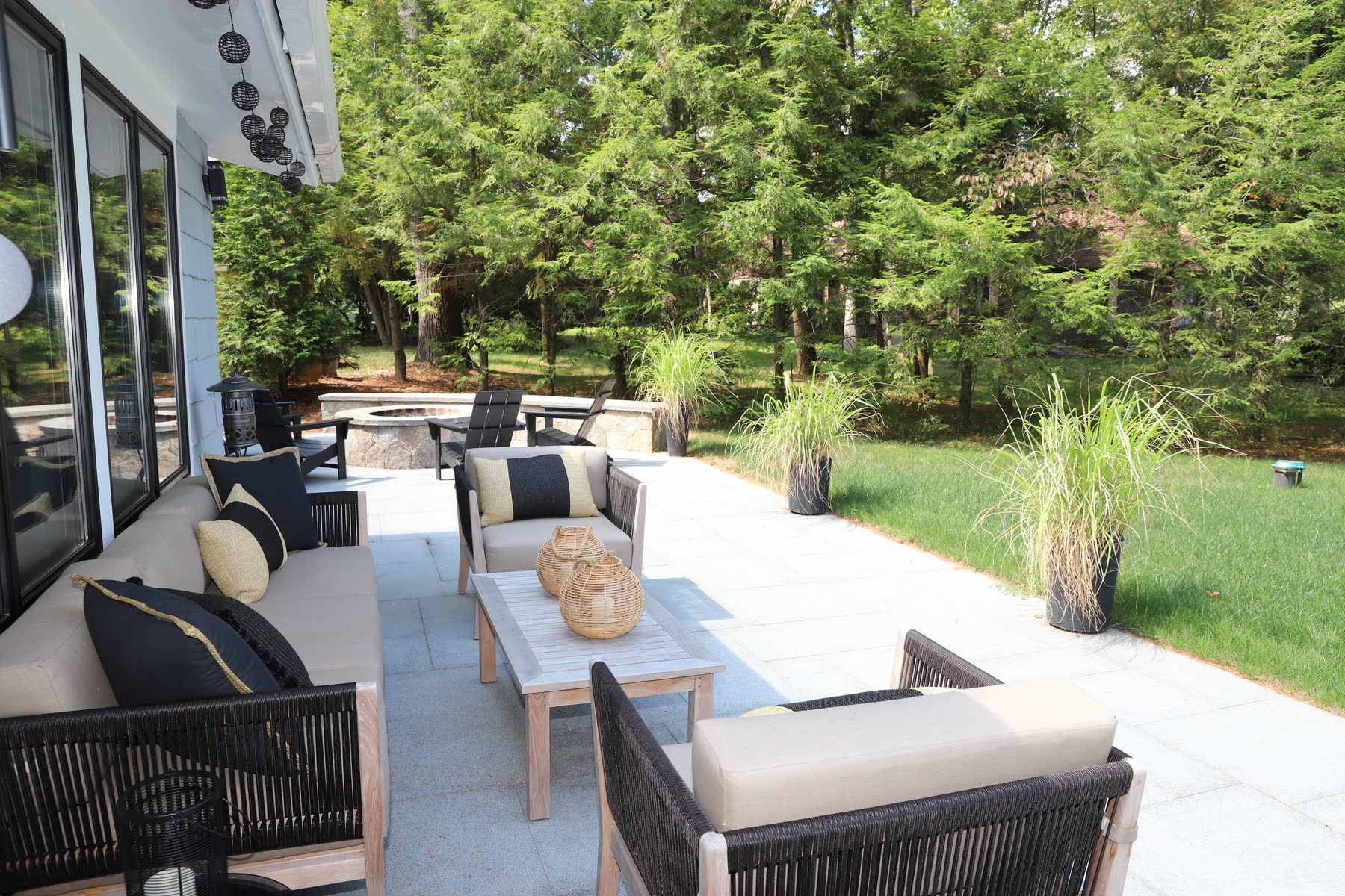 Bluestone patio at Sunset Drive with lounge seating and a fire-pit area opening onto the lawn and pines — Bulldog Enterprises.