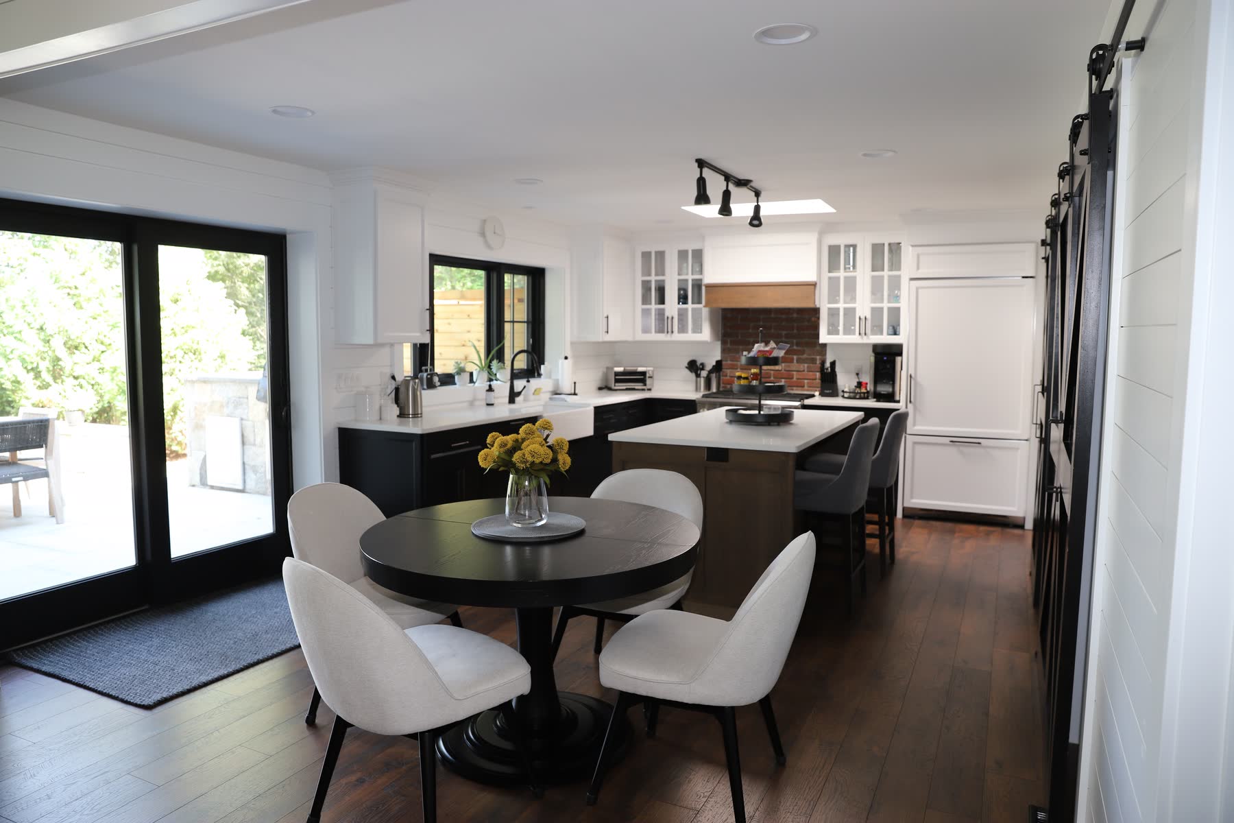 Open kitchen and dining at Sunset Drive with white cabinetry, a dark island, a brick backsplash and sliding doors to the patio — Bulldog Enterprises.
